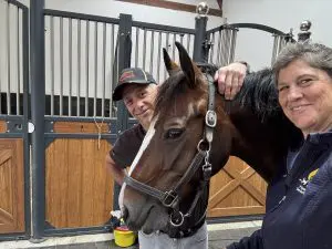 Andy Marcoux in a Coachman's Delight cap, the bay mare Loretta, and Tara Devine together in the stable aisle at Genderstyne Stables in Steensel, Netherlands."