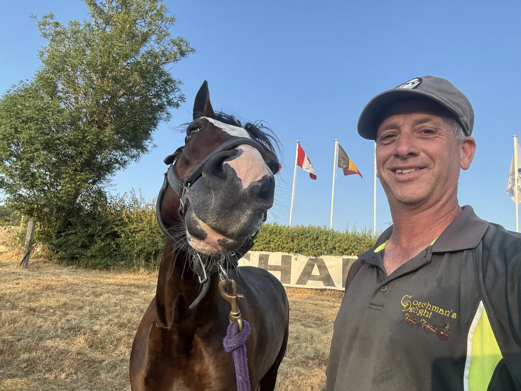 Andy Marcoux in a Coachman's Delight polo grinning beside Loretta, the bay mare pulling a goofy face toward the camera, national flags flying behind them at the Chablis CDE.