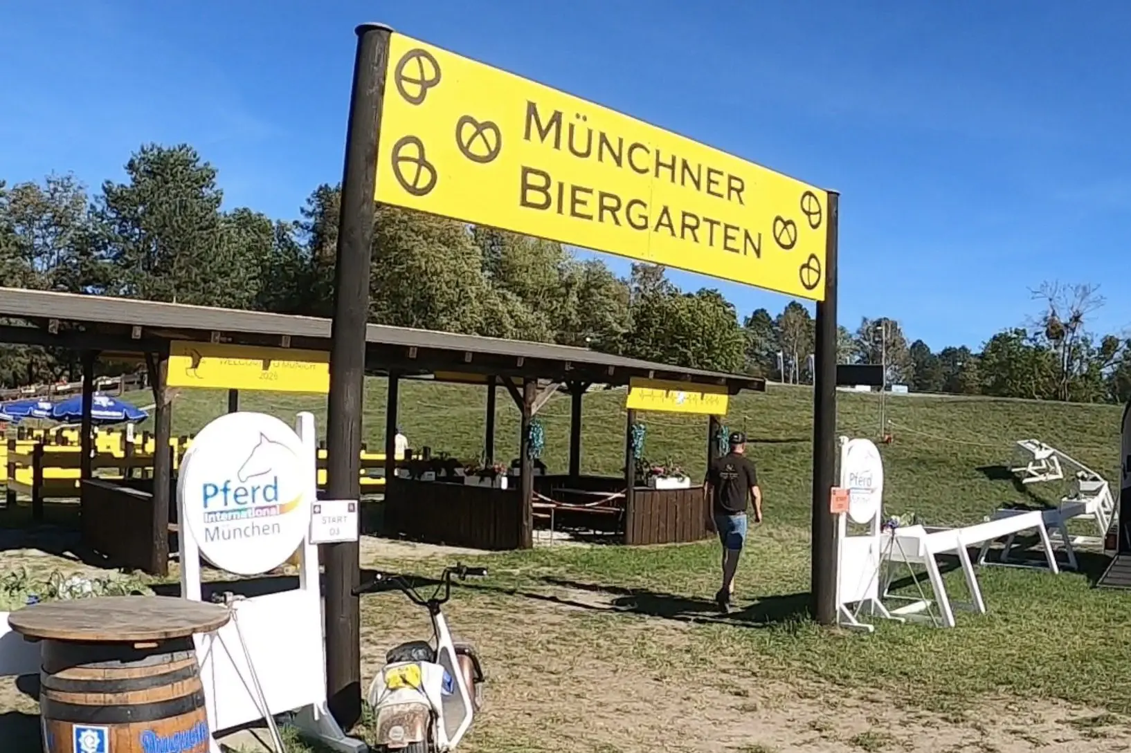 The big yellow Münchner Biergarten sign with pretzel logos over the start of Marathon Obstacle 3, with a beer barrel table and the obstacle's wooden beer hut behind, at the 2026 FEI Driving World Championship for Single Horses in Munich.